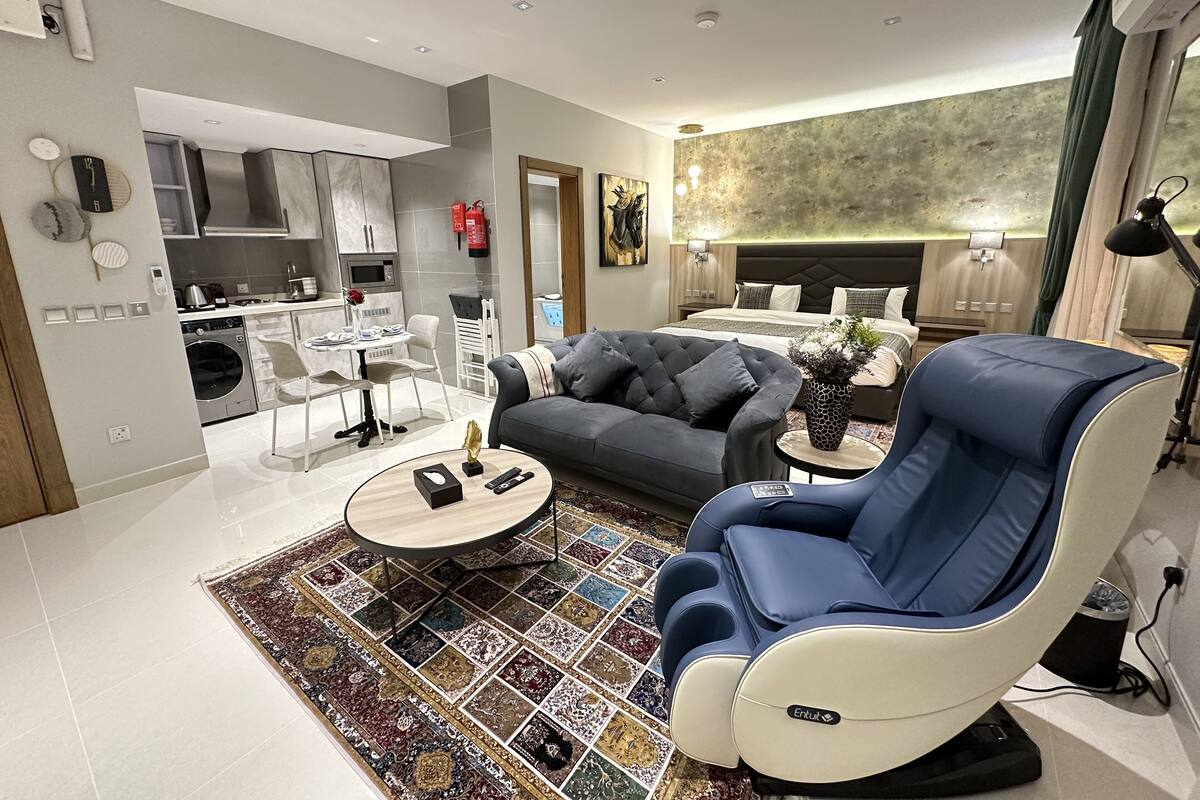 A modern living room with a grey sofa, a blue recliner, a coffee table, and a patterned rug. A kitchen with white cabinets and a dining area with a table and chairs are visible in the background.