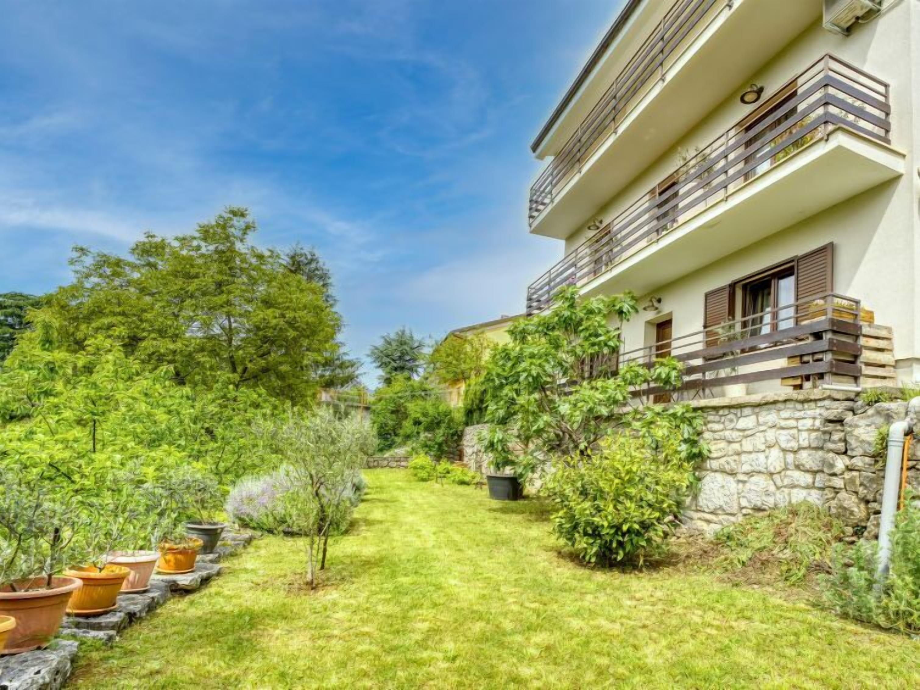 A two-story building with a stone wall and a balcony. There is a well-maintained lawn with potted plants and trees.