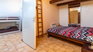 Een slaapkamer met een stapelbed, een trapje, een bed met een beddeken met patroon, een radiator en een houten plafondbalk.