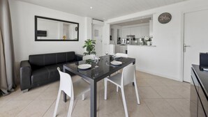 A modern living room with a black sofa, a glass dining table with white chairs, a potted plant, and a kitchen visible through an open door. - Medusas by Interhome (Dénia)