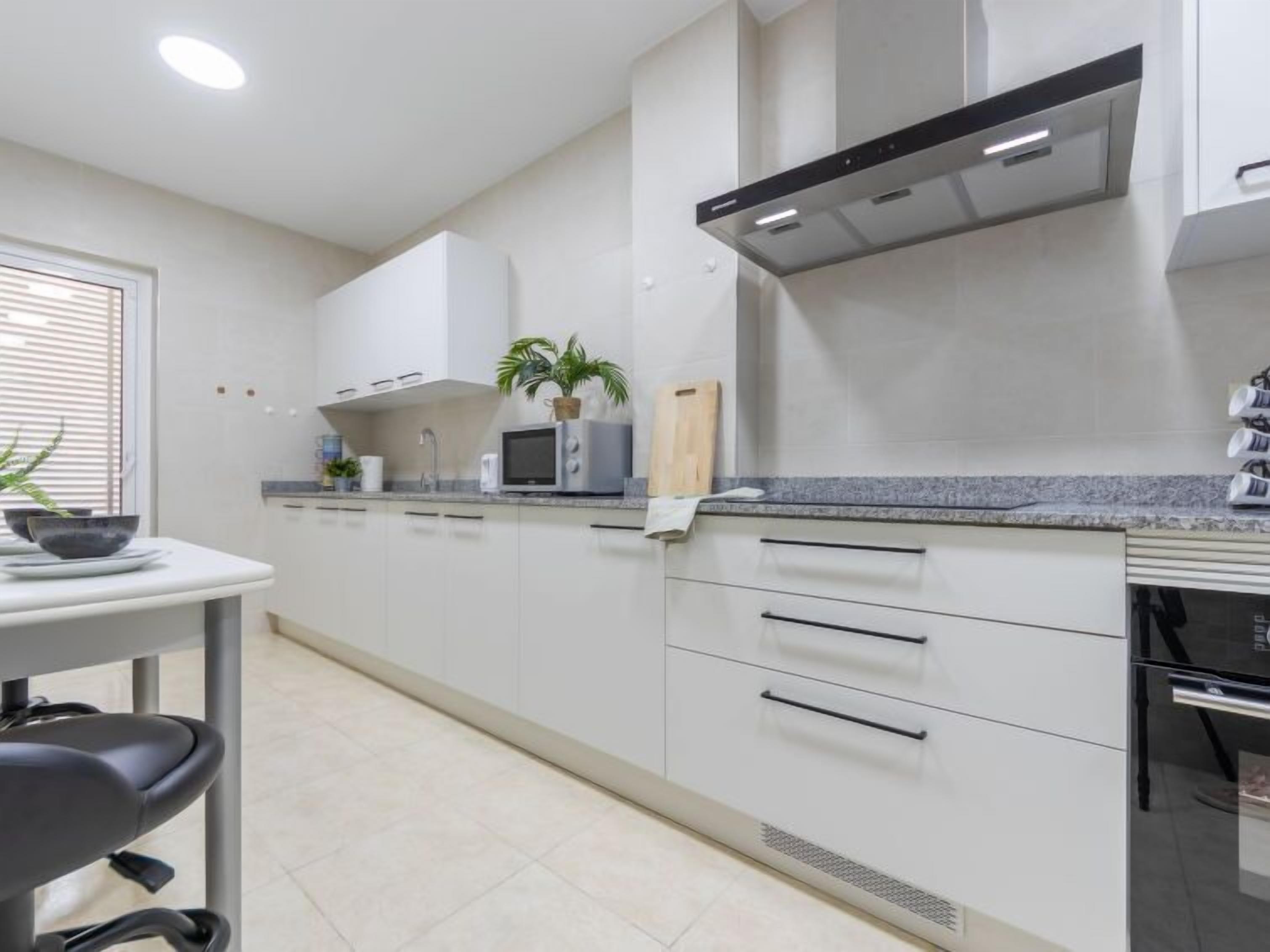 A modern kitchen with white cabinets, a microwave, a plant, and a window with blinds.
