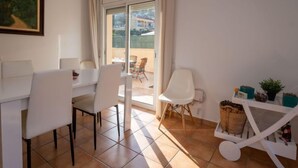 Un espace repas moderne avec une table et des chaises blanches, une petite table d’appoint blanche avec un panier, et une vue sur un balcon avec une chaise.