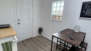 Una habitación con piso de madera, una puerta blanca, un ventanal pequeño, una mesa con sillas y un ventilador de techo.
