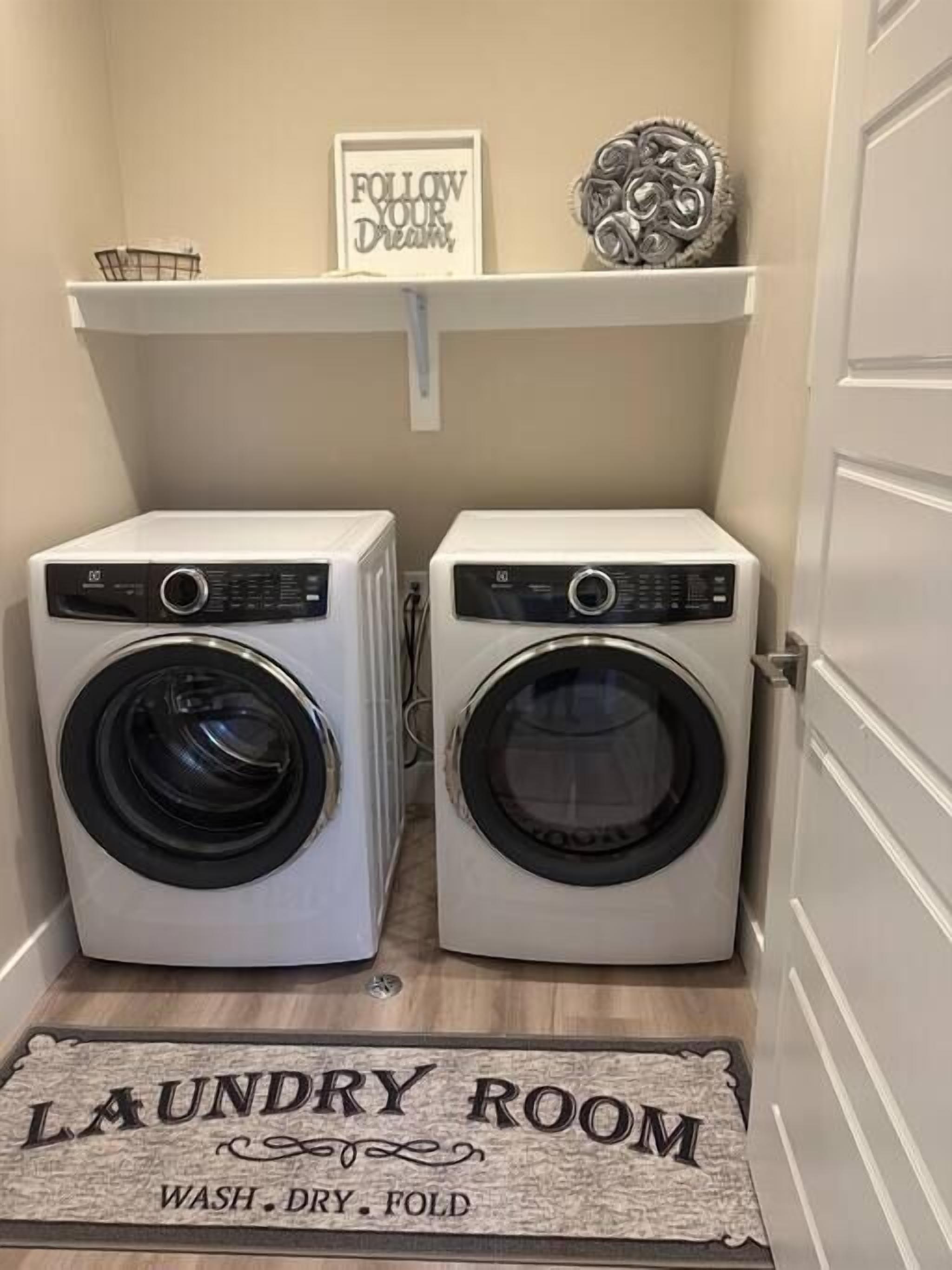 A laundry room with two front-loading washing machines, a shelf with a motivational sign, and a doormat that reads "LAUNDRY ROOM WASH. DRY. FOLD."