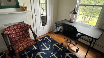Een kamer met een houten vloer, een bureau met een lamp, een stoel, een deur met een spiegel en een raam met uitzicht op groen.