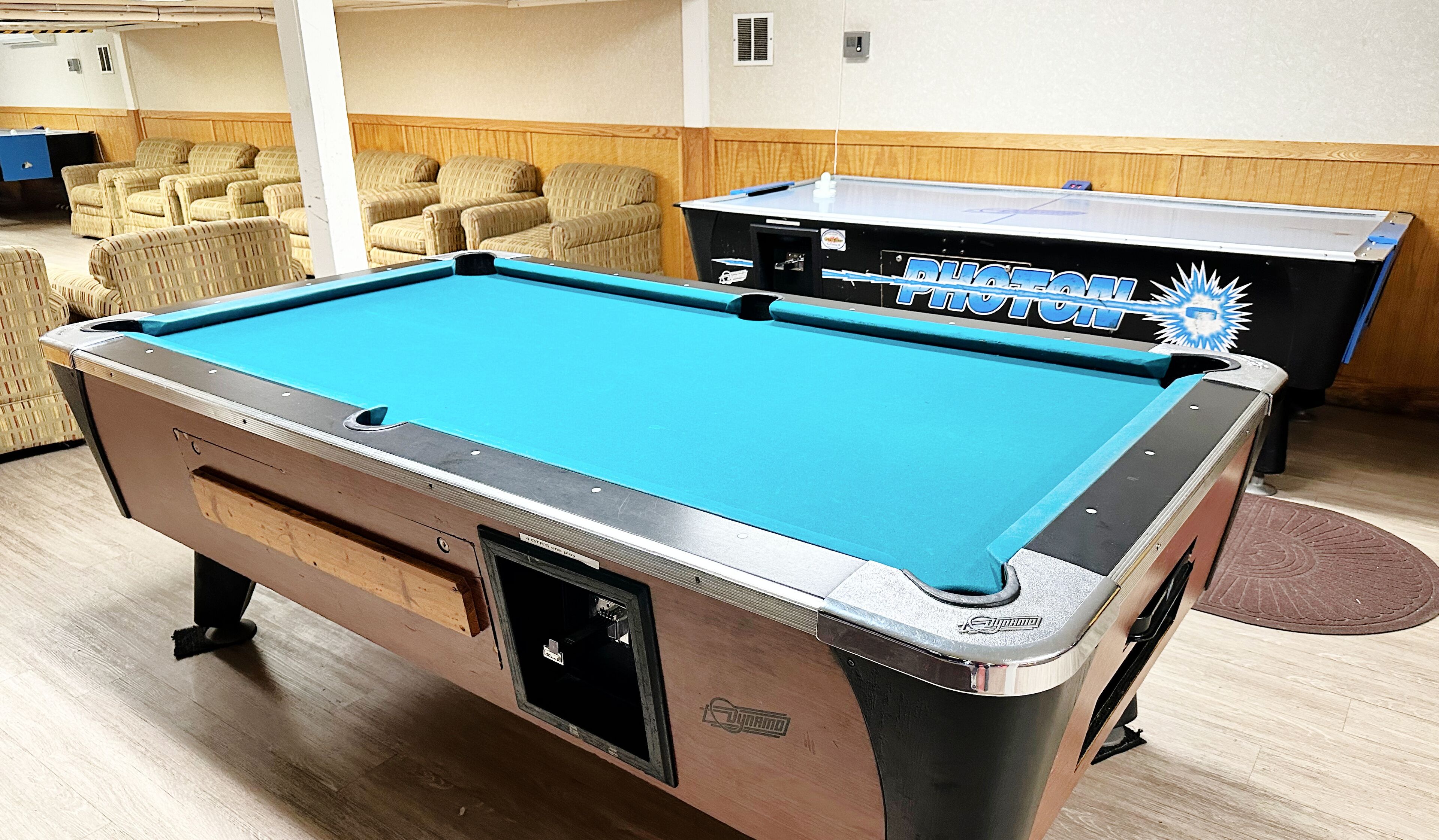 Una mesa de billar con superficie azul y una mesa de ping pong con red, en una sala con paneles de madera y muebles de mimbre.