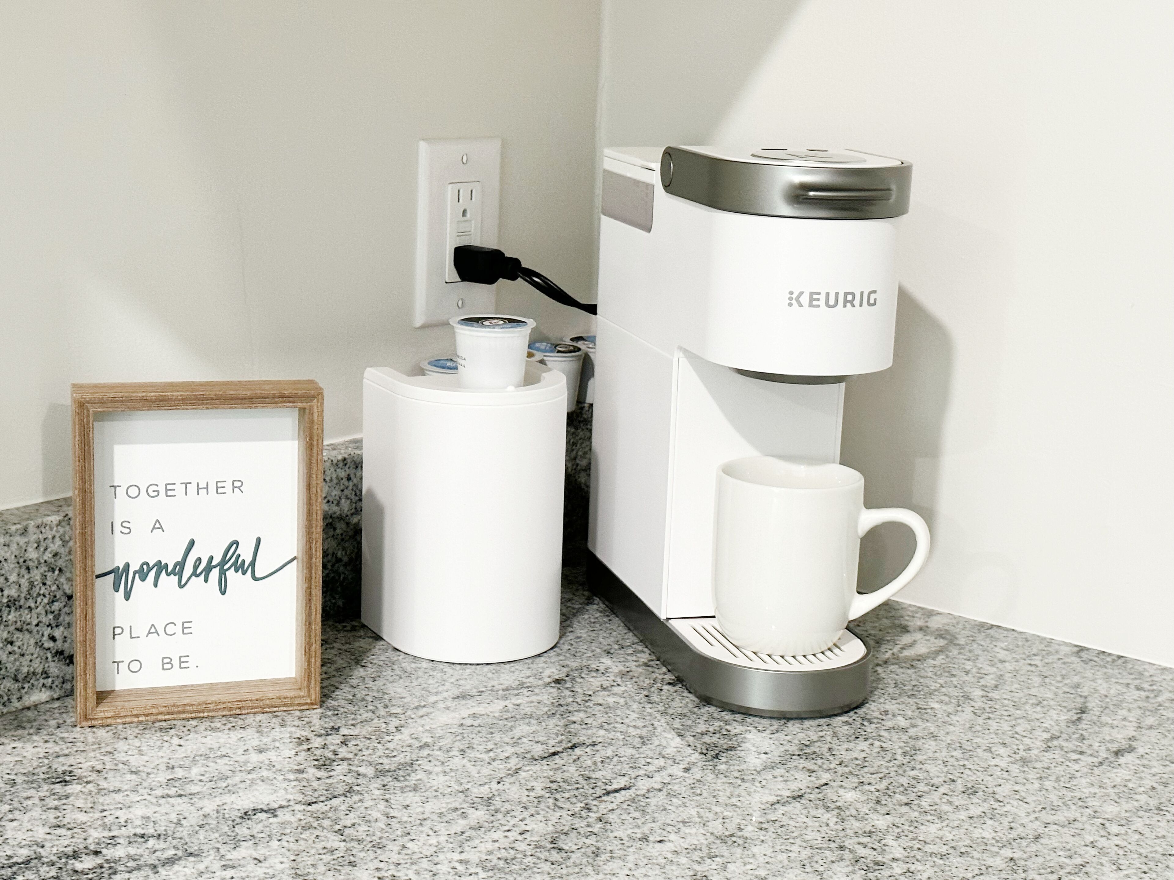 Eine Kaffeemaschine mit Tasse auf der Arbeitsplatte, ein gerahmter Spruch und ein Stecker in der Wand.