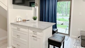 Une cuisine moderne avec des meubles blancs, un plan de travail en granit, un téléviseur noir fixé au-dessus du plan de travail, une plante en pot et un rideau bleu.