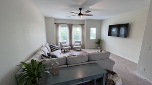 A modern living room with a sectional sofa, two armchairs, a coffee table, a ceiling fan, and a TV mounted on the wall.