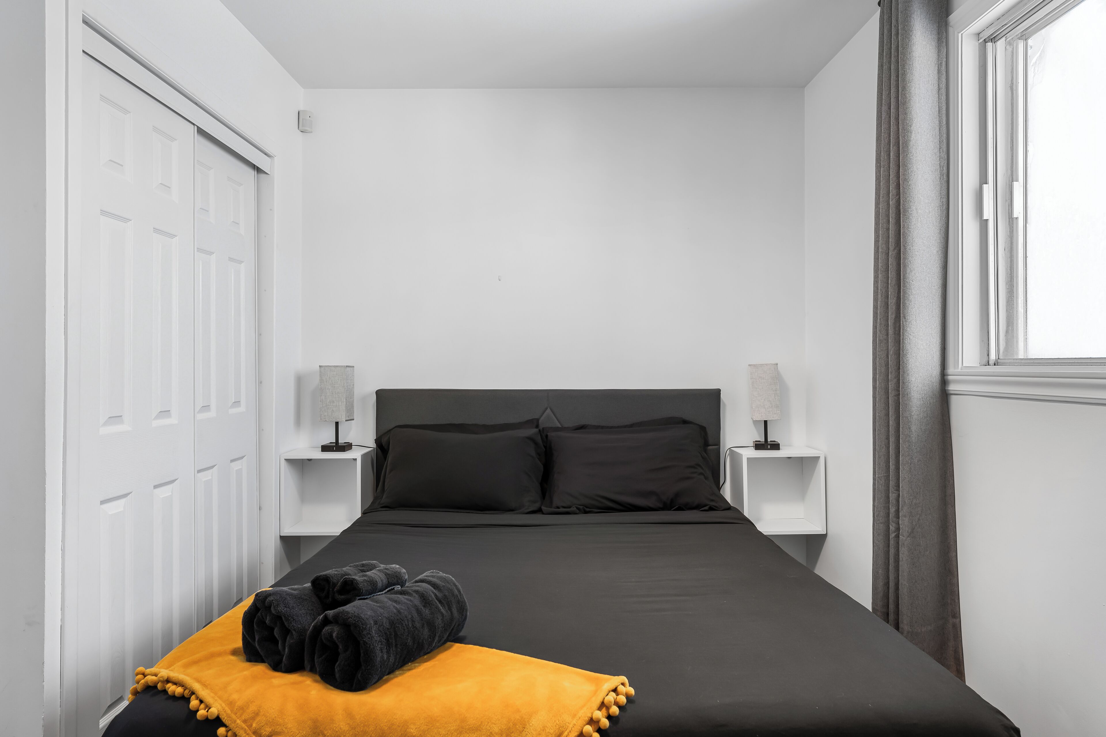 Un dormitorio con una cama negra, mesitas de noche blancas, un cabecero negro y una manta amarilla.