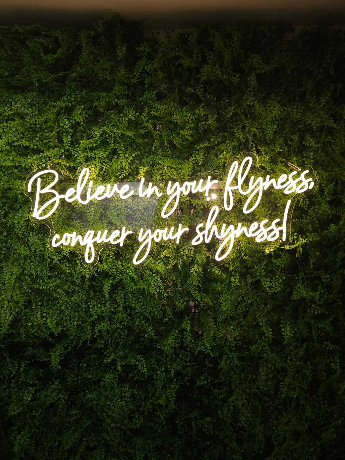 Un letrero de neón con el texto 'Believe in your flyness, conquer your shyness!' sobre un fondo de follaje verde exuberante.