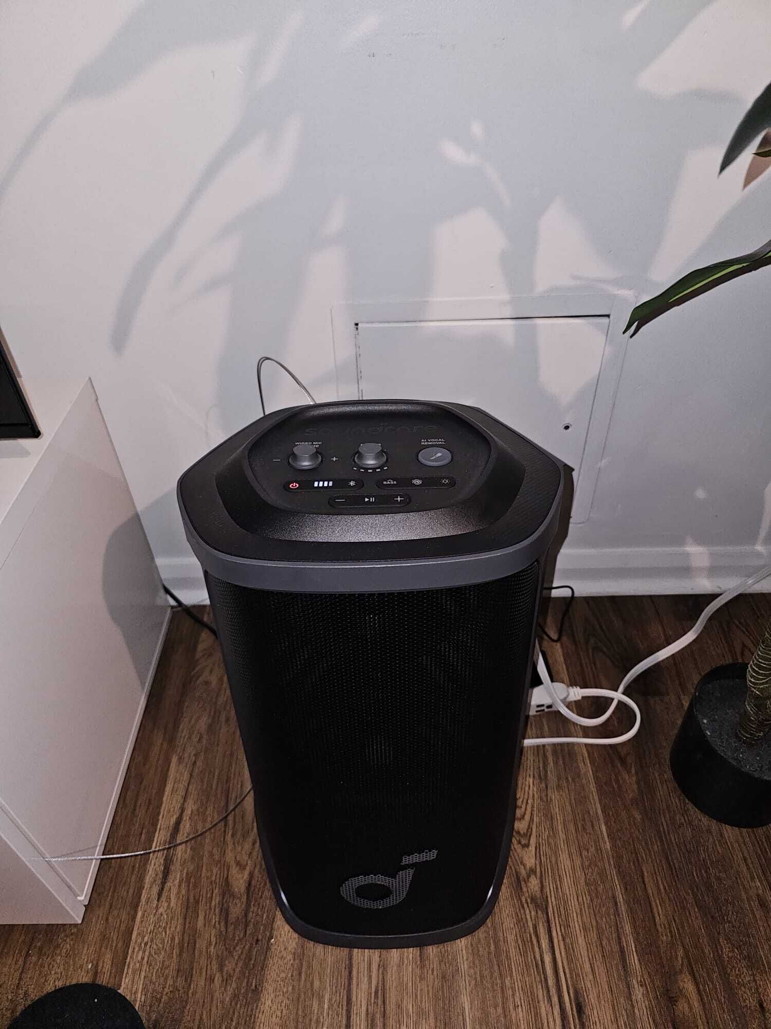 A black portable speaker with a control panel, placed on a wooden floor against a white wall.