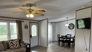 A living room with a sofa, coffee table, dining table, chairs, a ceiling fan, and a TV mounted on the wall. - Kim's Place (Cookeville)