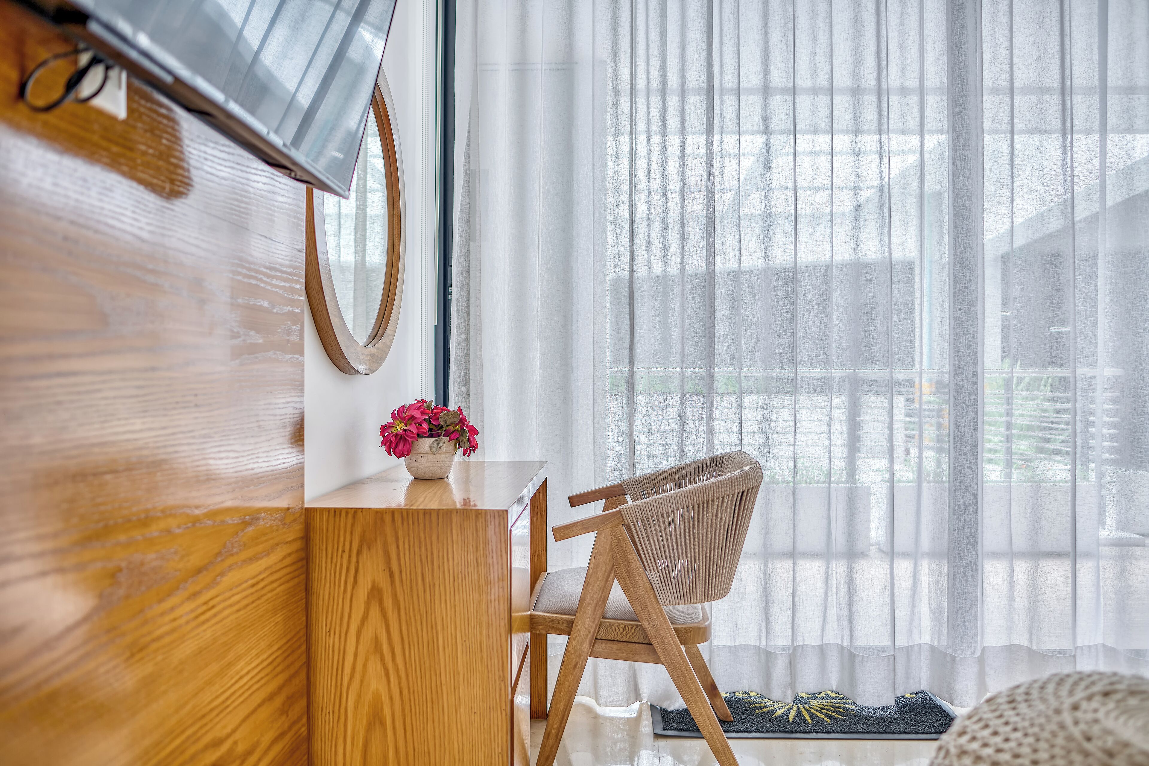 A wooden desk with a vase of flowers, a chair, and a large window with sheer curtains.
