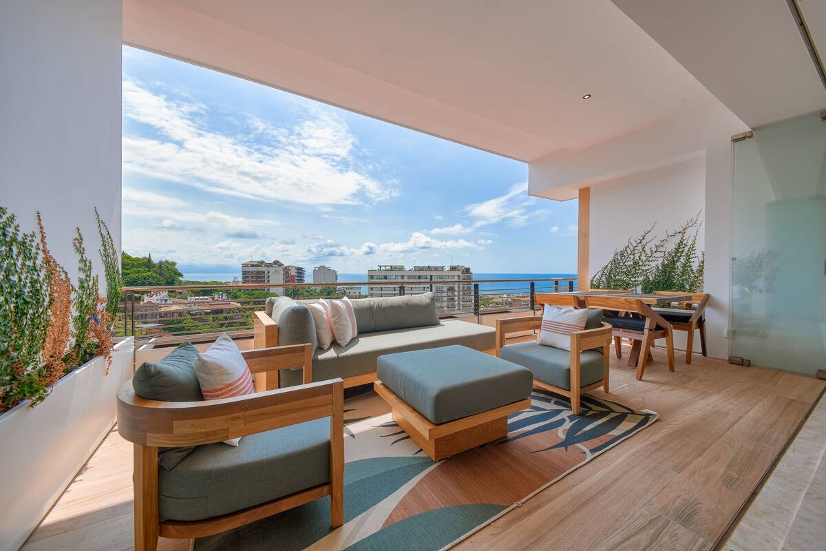 Un balcon moderne avec un plancher en bois, des canapés gris, des chaises en bois et une table en verre.