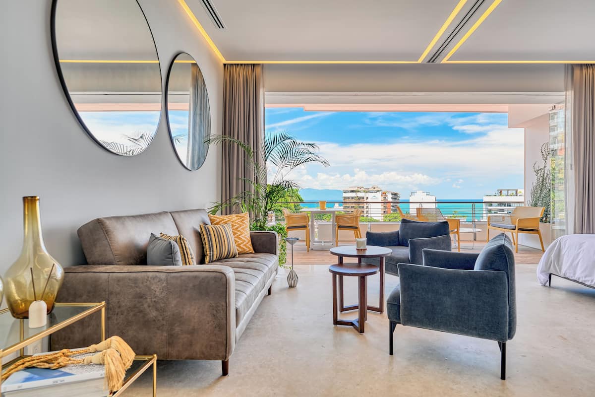 Une chambre d’hôtel moderne dotée d’un canapé, de fauteuils, d’une table basse et d’un balcon offrant une vue sur l’océan et les bâtiments.