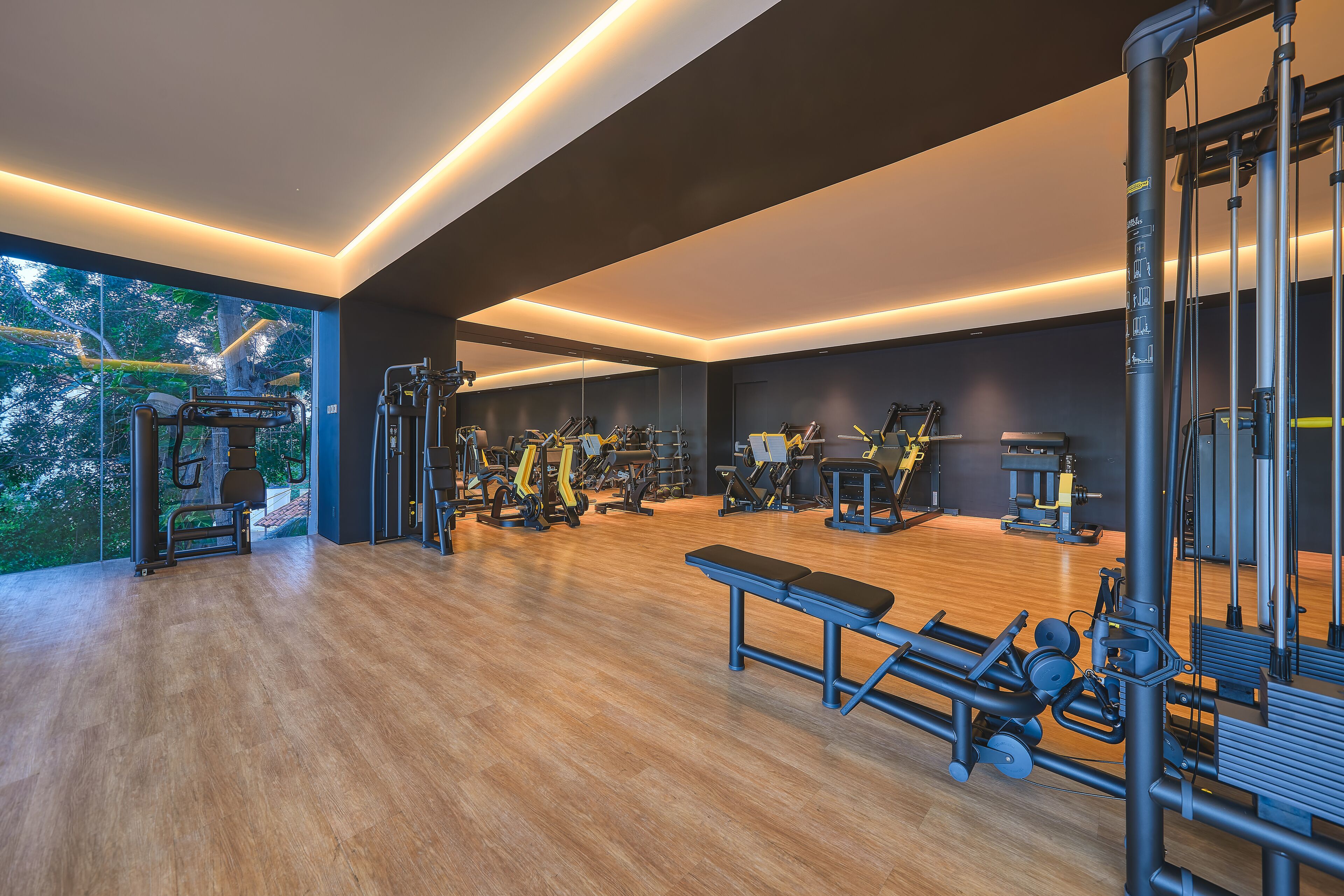 Un gimnasio moderno con diversas máquinas de ejercicio, piso de madera y amplias ventanas con vistas a árboles.