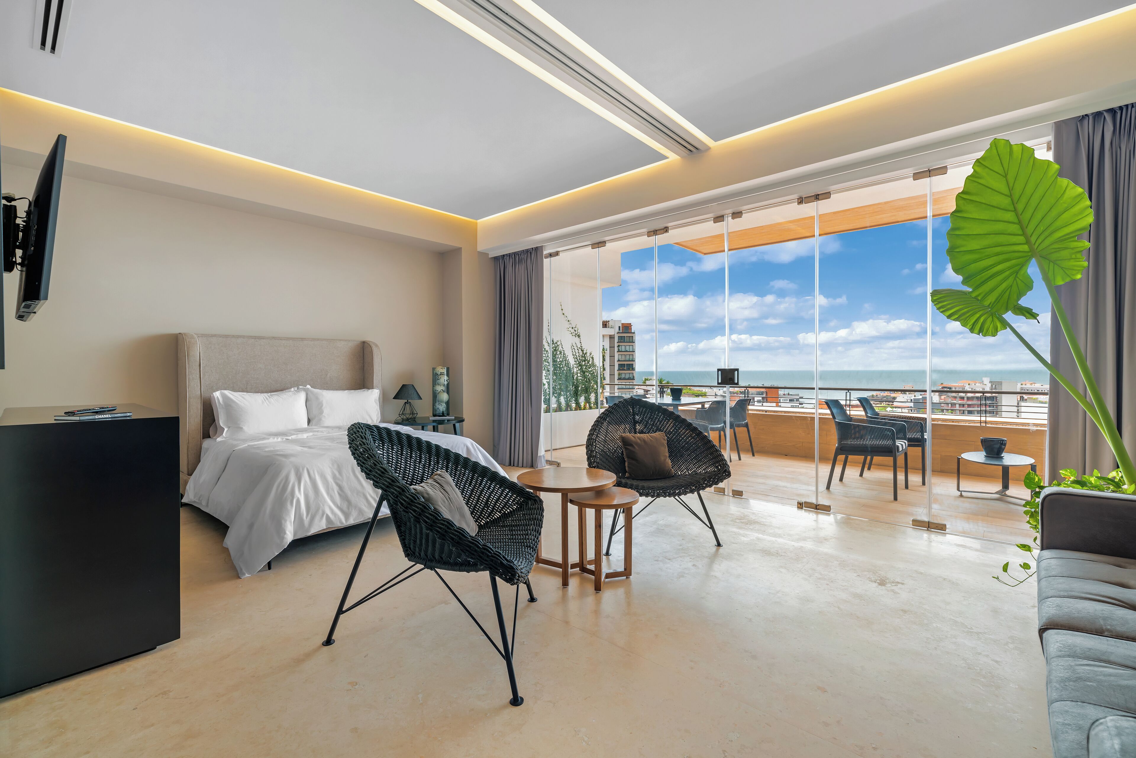 Une chambre d’hôtel moderne équipée d’un lit, de deux fauteuils, d’une petite table et d’une grande fenêtre donnant sur la ville et l’océan.