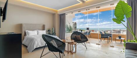 Une chambre d’hôtel moderne équipée d’un lit, de deux fauteuils, d’une petite table et d’une grande fenêtre donnant sur la ville et l’océan.