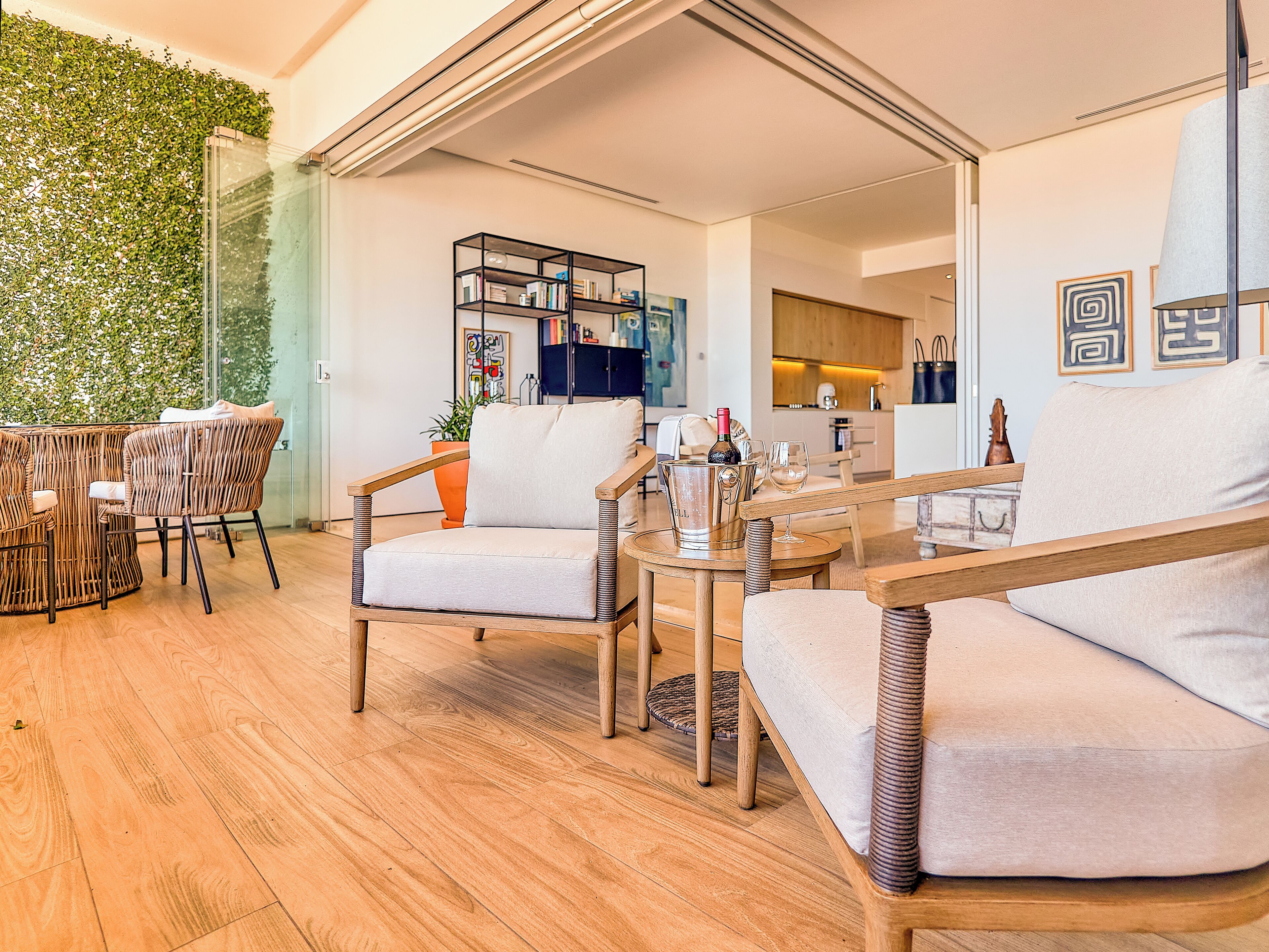 A modern living room with a glass wall, a wooden floor, and a dining area with wicker furniture.
