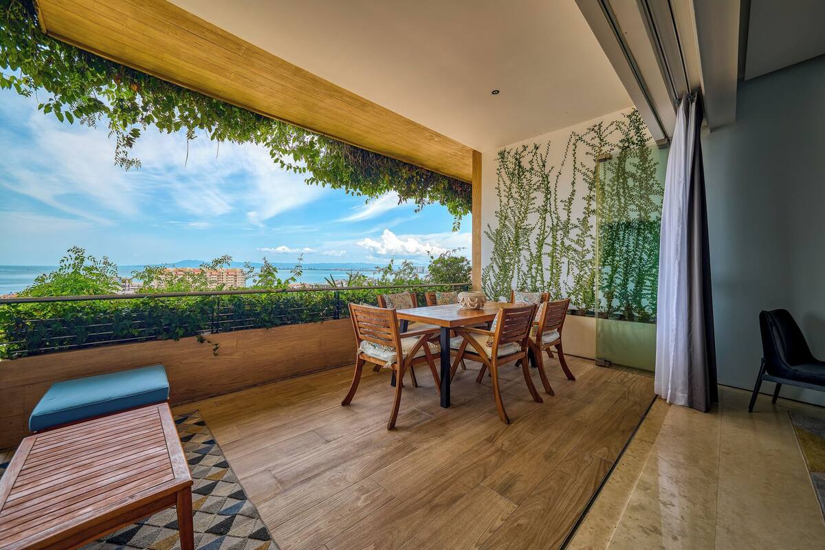 Un balcon avec une table et des chaises en bois, un banc bleu et une vue sur la mer et les bâtiments.