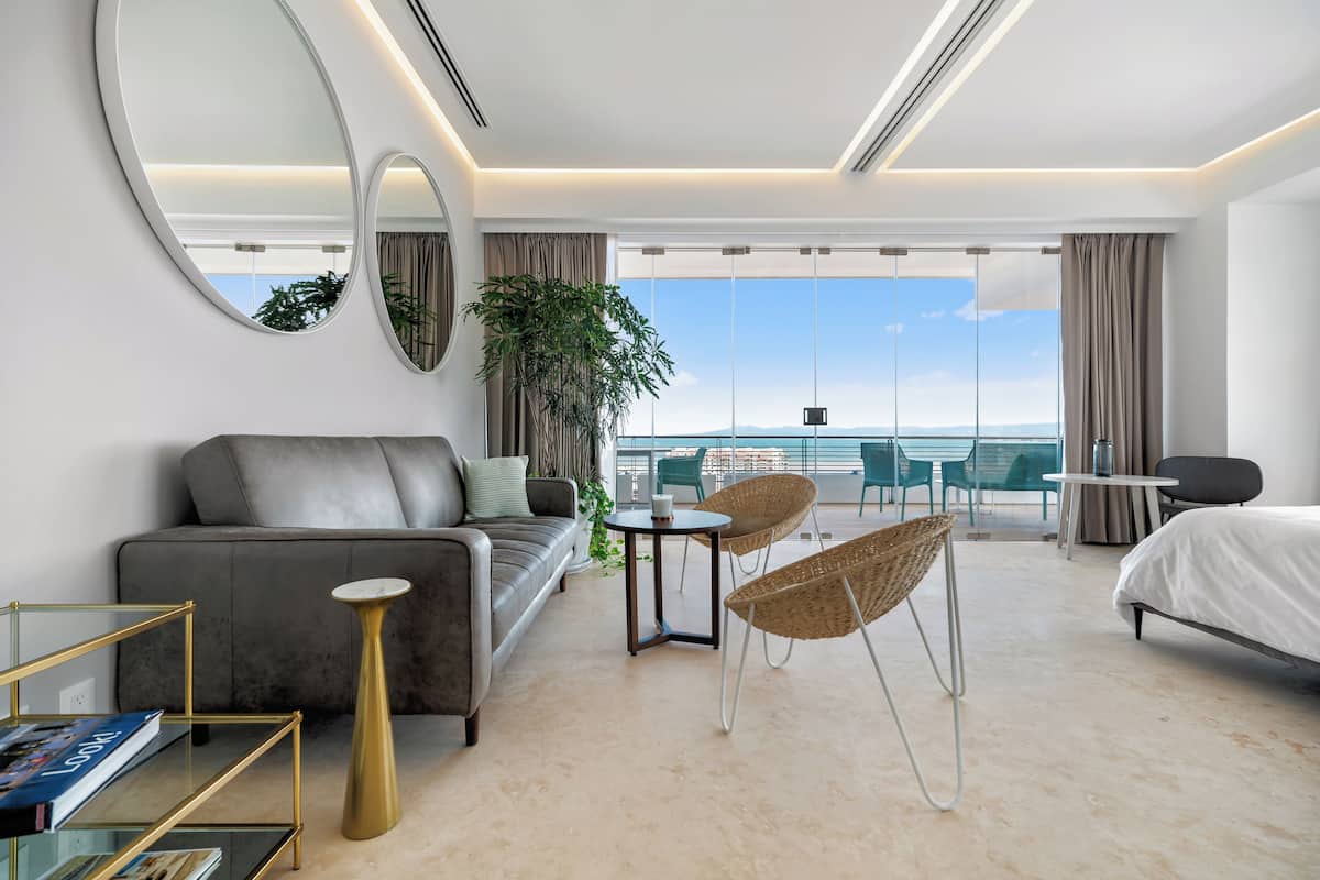Une chambre d’hôtel moderne avec un canapé, un miroir rond, une petite table, une chaise en osier, un lit et un balcon avec vue.