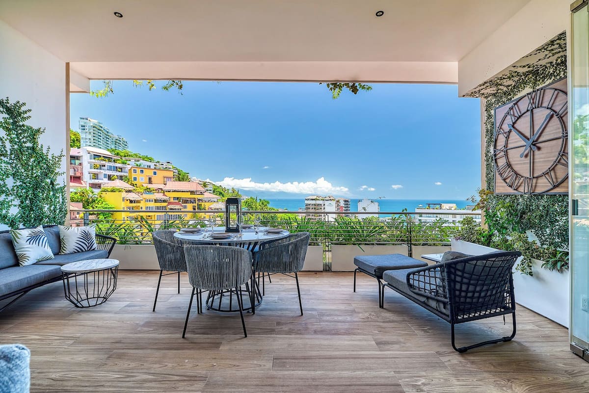 Un balcon avec une table ronde et des chaises, un canapé, et une vue sur la mer et les bâtiments.