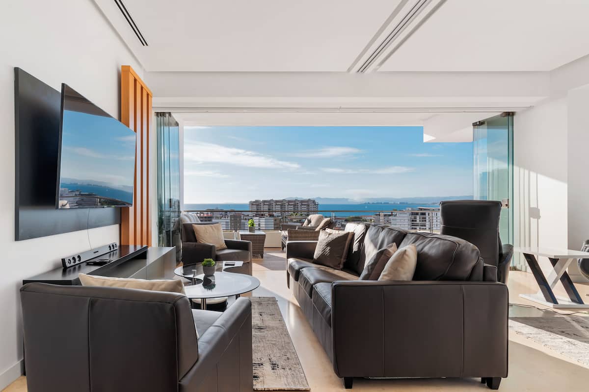 Un salon moderne doté d’un grand téléviseur à écran plat, de canapés en cuir, d’une table basse et offrant une vue sur le paysage urbain et l’océan.