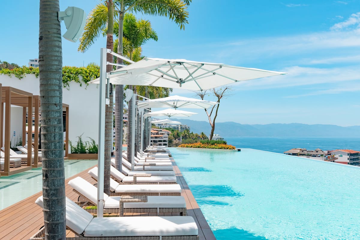 Un espace aménagé au bord de la piscine, avec des chaises longues et des parasols, une terrasse en bois et une vue sur l’océan.