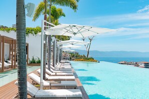 Un espace aménagé au bord de la piscine, avec des chaises longues et des parasols, une terrasse en bois et une vue sur l’océan.