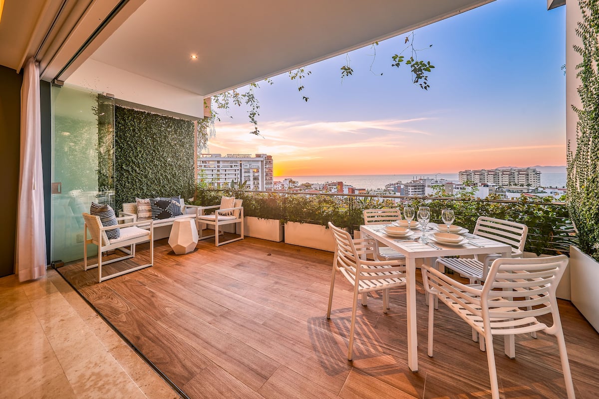 Un balcon doté d’un plancher en bois, de meubles blancs et offrant une vue sur le coucher de soleil sur la mer.