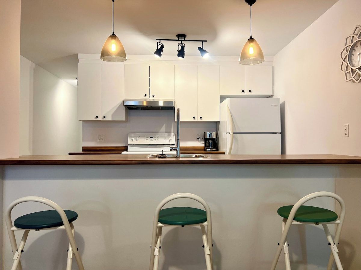 A kitchen with white cabinets, a stainless steel range hood, a white refrigerator, and two bar stools with green cushions.