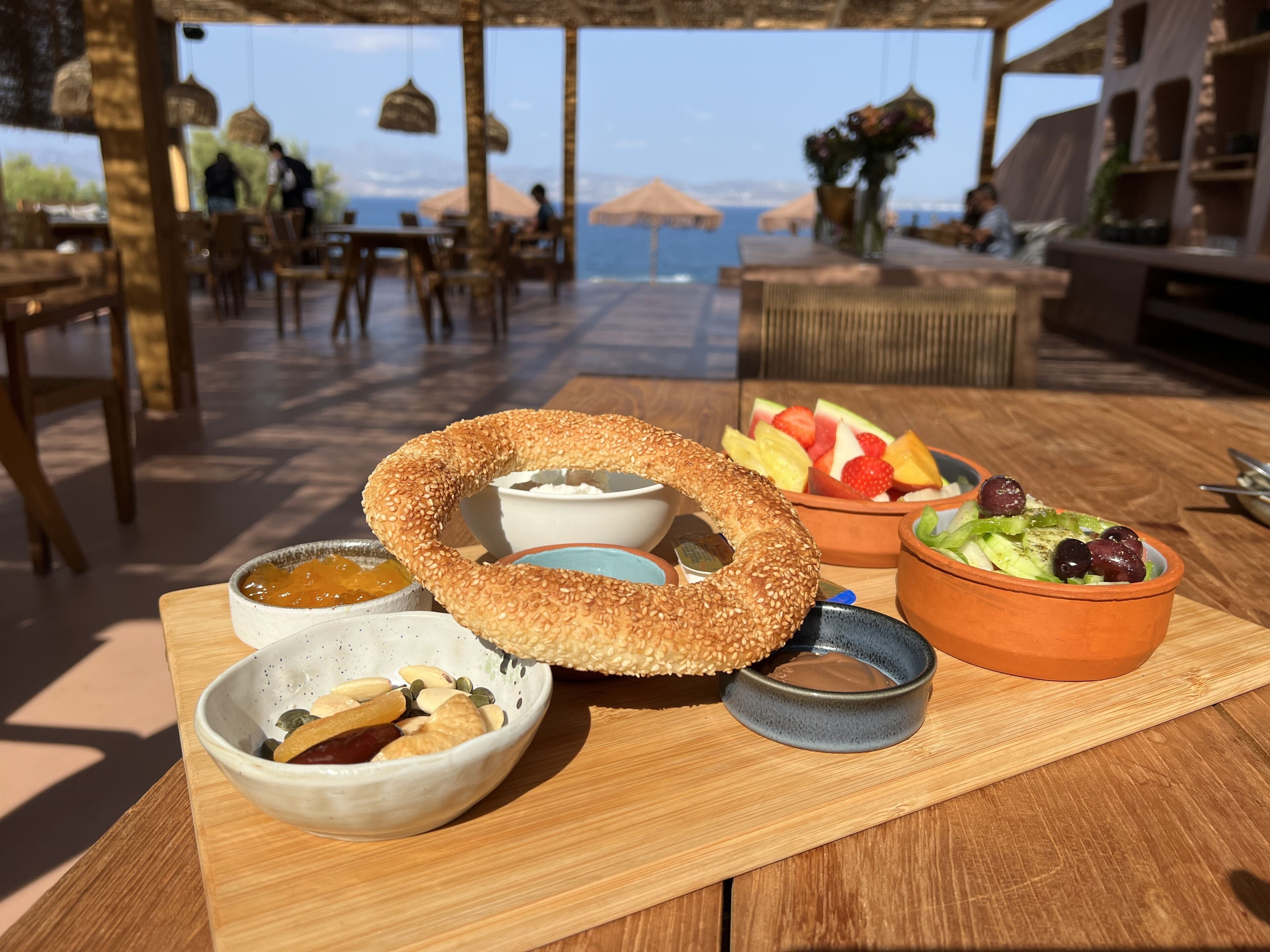 Una mesa con un bagel grande de semillas de sésamo, tazones de frutos secos y salsas, un bol de fruta y una ensalada pequeña.