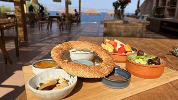 Una mesa con un bagel grande de semillas de sésamo, tazones de frutos secos y salsas, un bol de fruta y una ensalada pequeña.