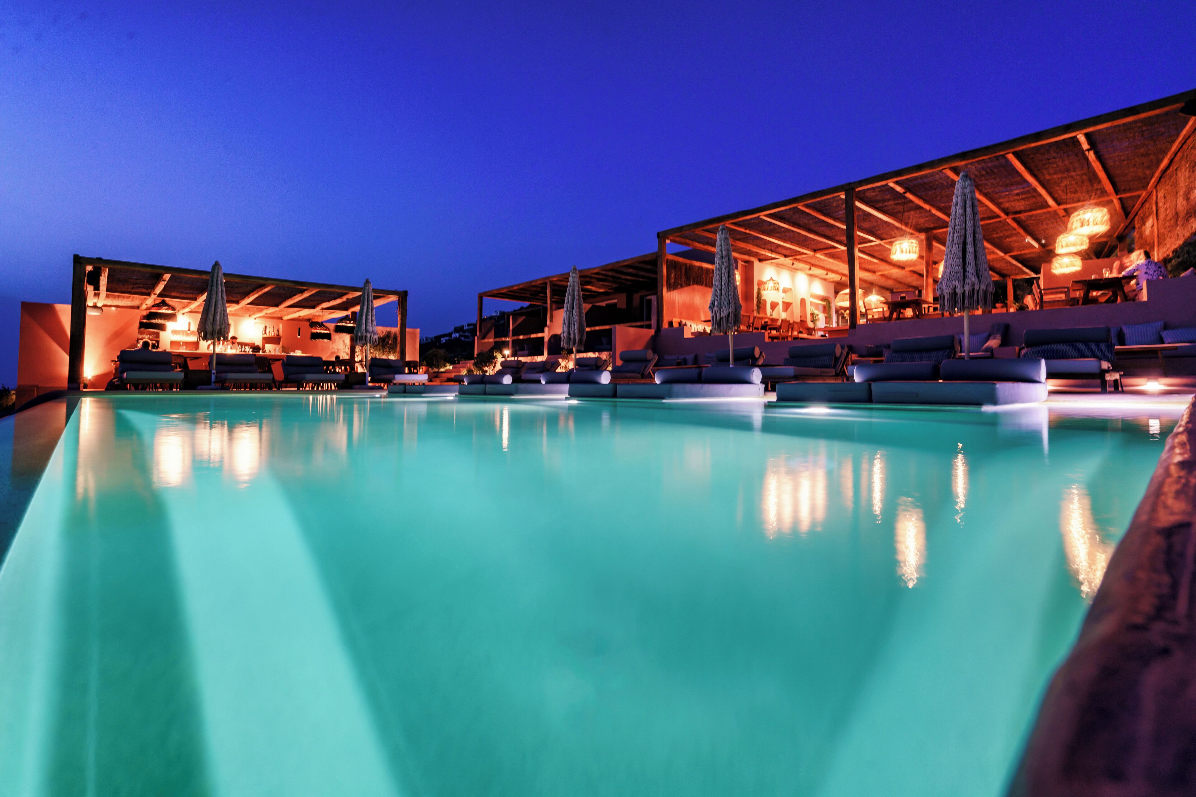 Un área junto a la piscina con sillones de descanso y sombrillas, un espacio cubierto con mesas y sillas, y una vista despejada de la piscina.