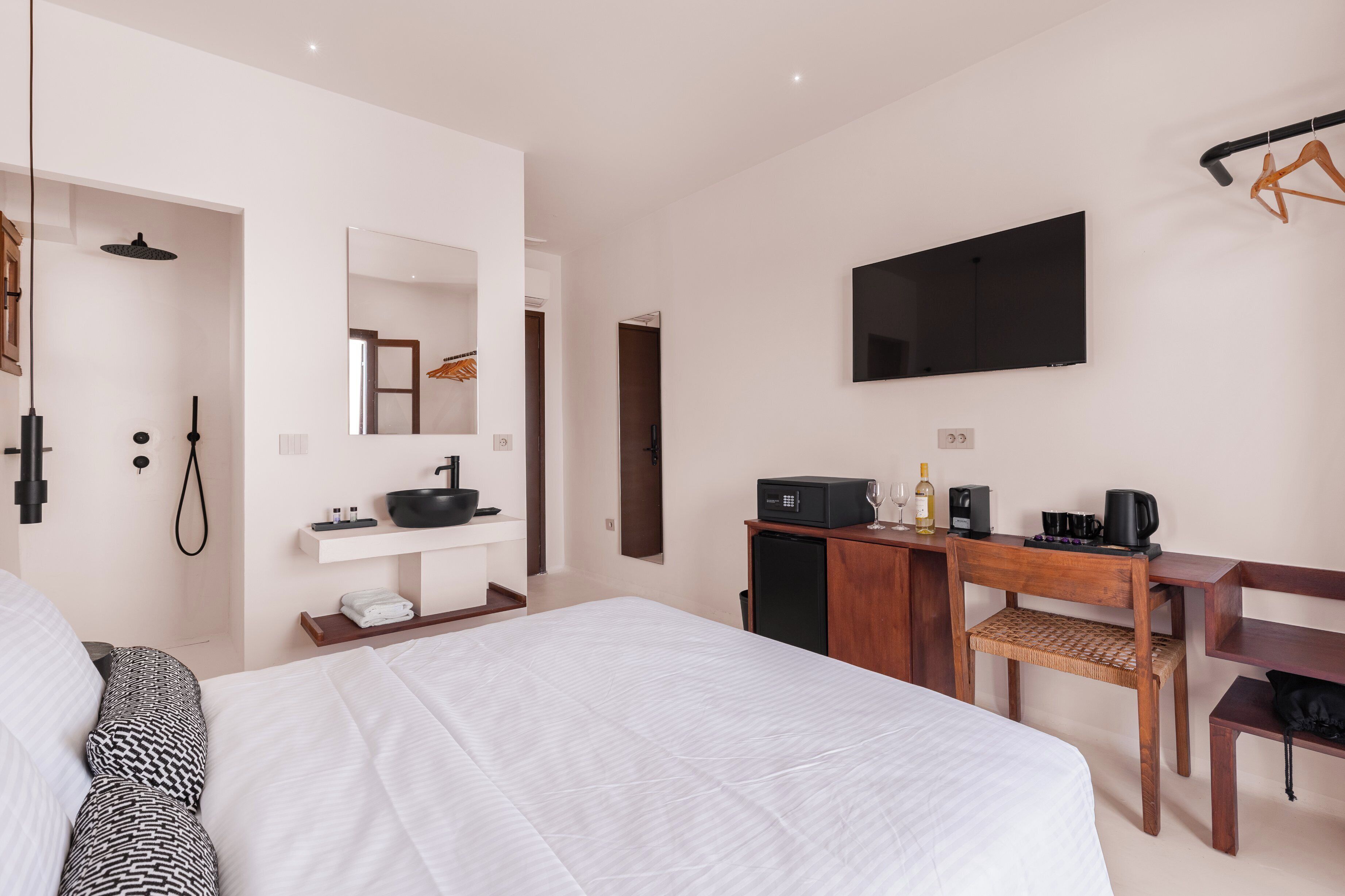 A modern hotel room with a large bed, a desk with a computer, a flat-screen TV, a mirror, and a small kitchenette.