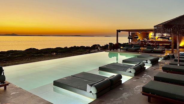 Une piscine à débordement avec des chaises longues, un espace bar et une vue sur la mer au coucher du soleil.