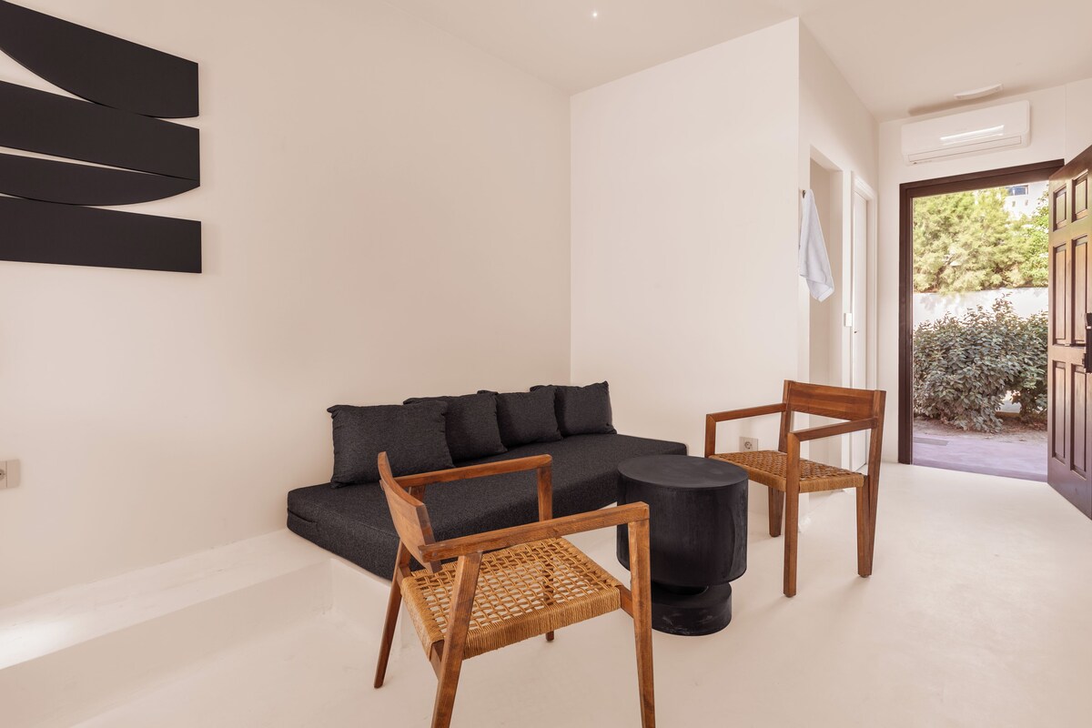 Uma sala de estar moderna com um sofá modular, duas cadeiras de madeira, uma mesa de centro redonda preta e uma obra de arte abstrata na parede.