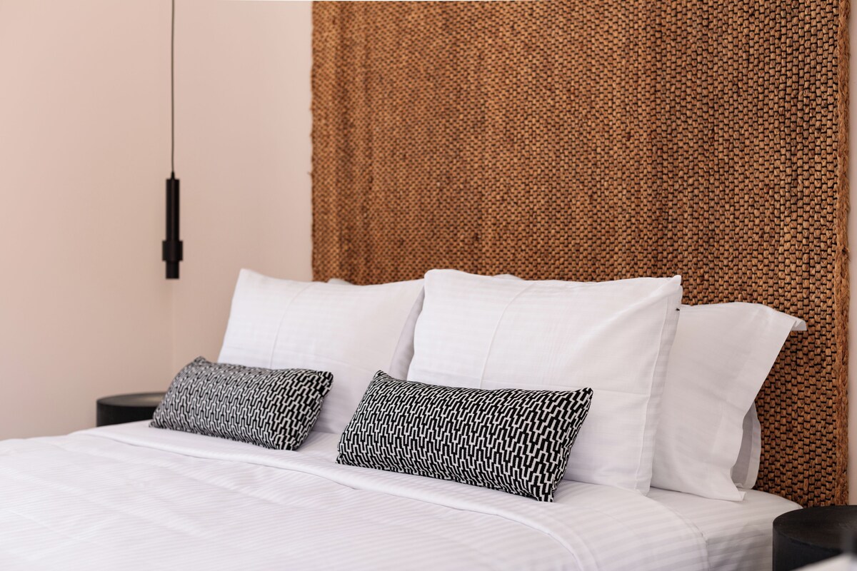 Cama bem arrumada com lençóis brancos e duas almofadas estampadas, cabeceira de fibra natural e abajur preto.