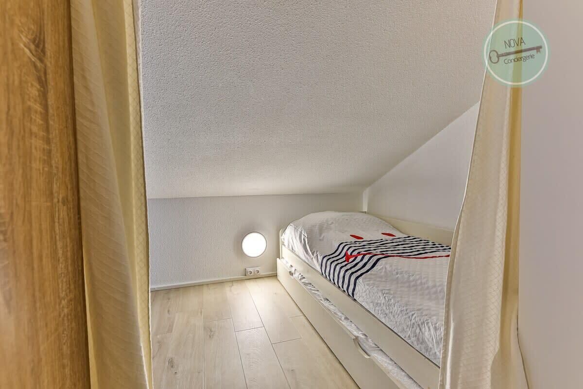 Une petite chambre avec un lit simple, un plafond en pente, un applique murale ronde et un lit recouvert d’une couverture à rayures.