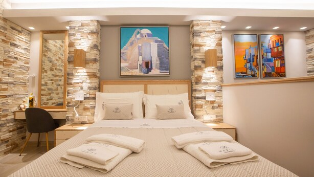 A hotel room with a large bed, two nightstands, a desk, and a chair. There are two paintings on the wall, a mirror, and a stone wall. - Mariet Athens Spa (Athens)