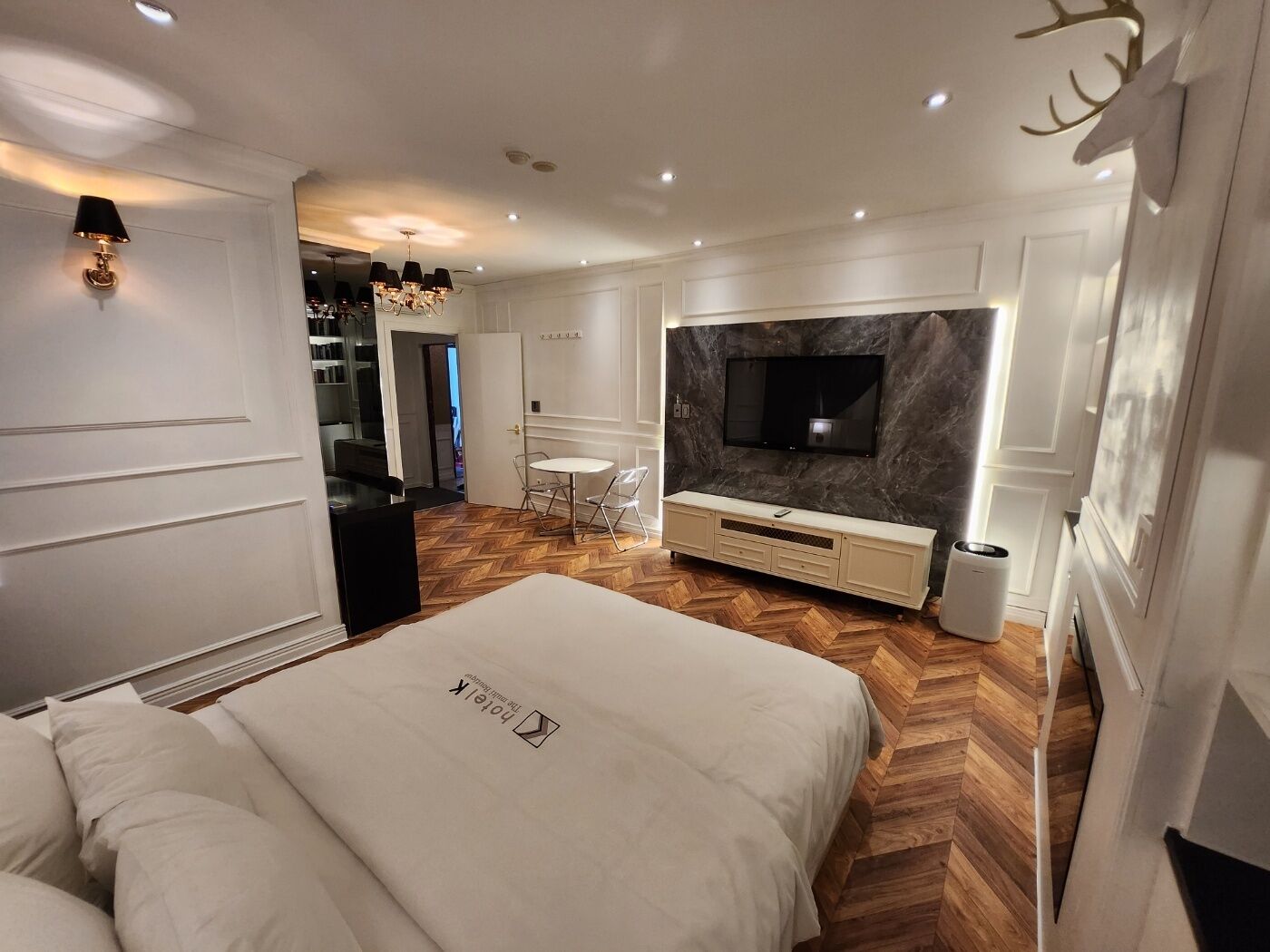 A modern bedroom with a large bed, a TV mounted on a marble wall, and a herringbone wood floor.