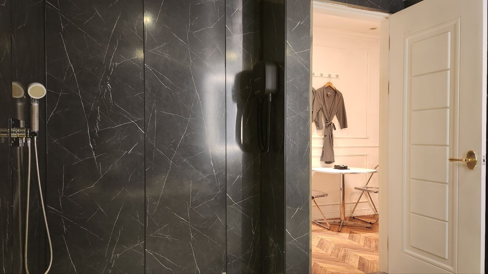 Une salle de bain avec un mur en marbre noir, un sèche-cheveux et une porte blanche donnant sur une pièce contenant une table et des chaises.