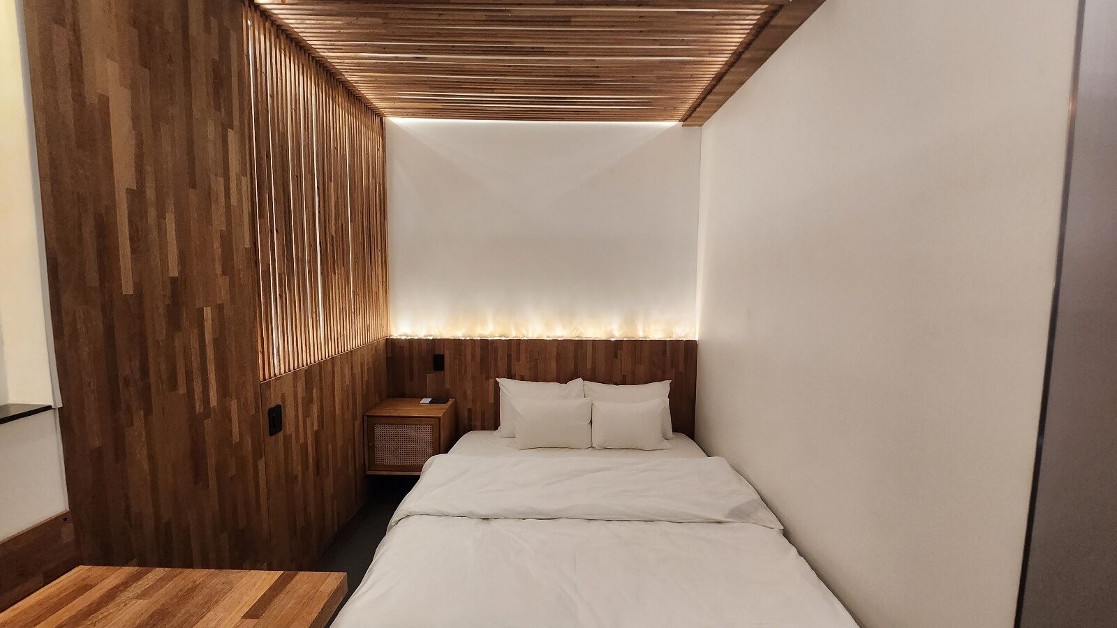 A compact room with a wooden slatted wall, a bed with white bedding, and a small wooden nightstand.