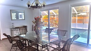A dining area with a glass table and wicker chairs, a vase with flowers, and a view of a garden through large windows. - Home with play room, enclosed pool . Only 10 minutes from the beach! (Vero Beach)