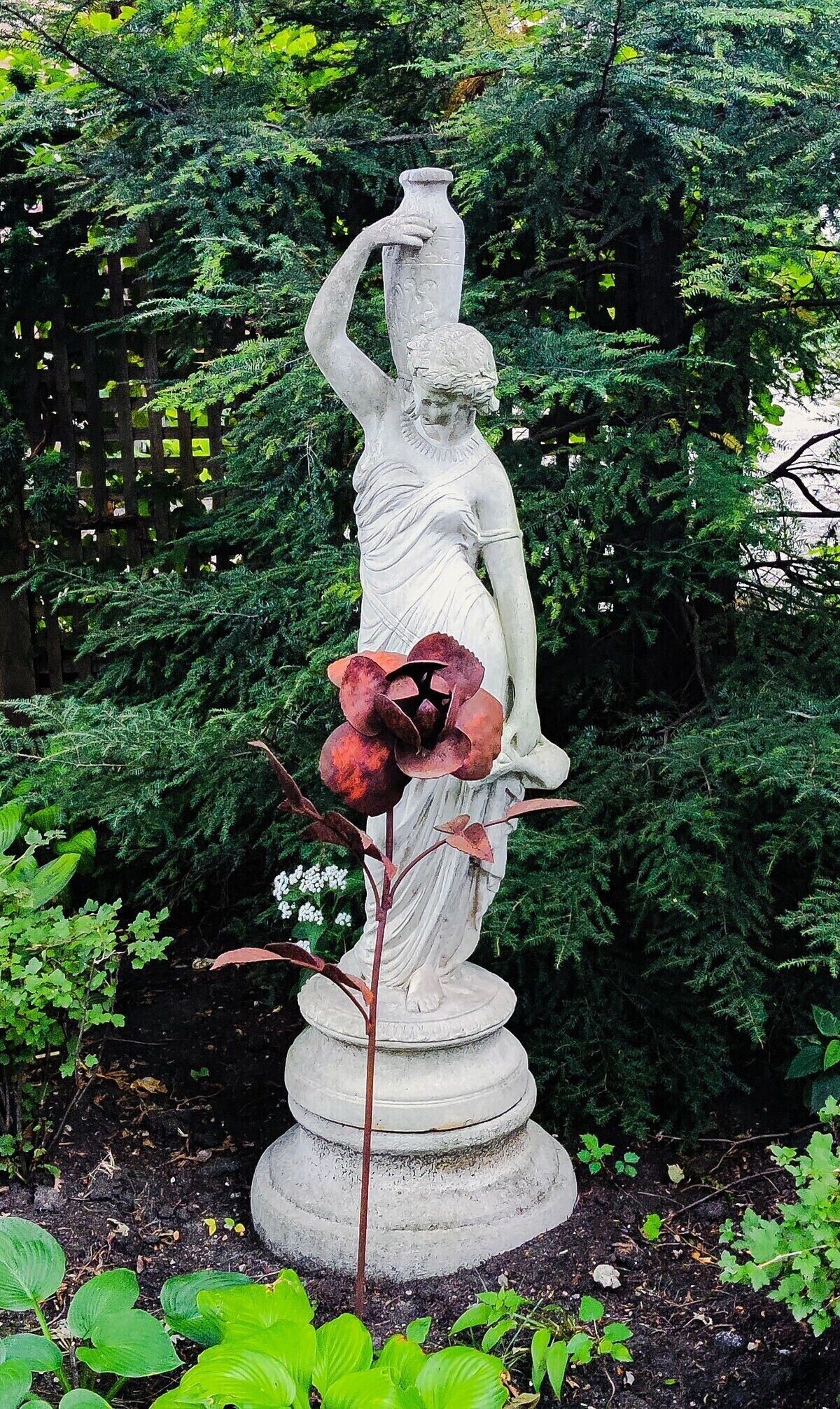 A statue of a woman holding a vase, surrounded by greenery and a wooden fence.