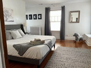 Quarto com cama grande, radiador, janela com cortinas, espelho e um elefante de pelúcia.
