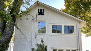 Ein zweistöckiges Haus mit Veranda, einer kleinen Terrasse mit Tisch und Stühlen und einem großen Baum im Vordergrund.
