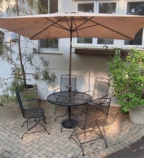 Um pátio com uma mesa redonda e quatro cadeiras, um guarda-sol e vasos com plantas.