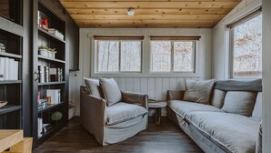 Une pièce chaleureuse avec un plafond en bois, deux fenêtres équipées de stores, une bibliothèque, un canapé et un fauteuil.
